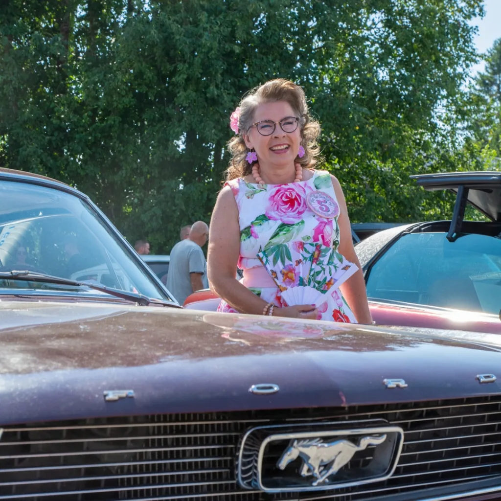 Nainen kukkamekossa vanhan jenkkiauton vieressä – talkoovoimin järjestettyjä elämyksiä ja yhteisöllistä iloa.