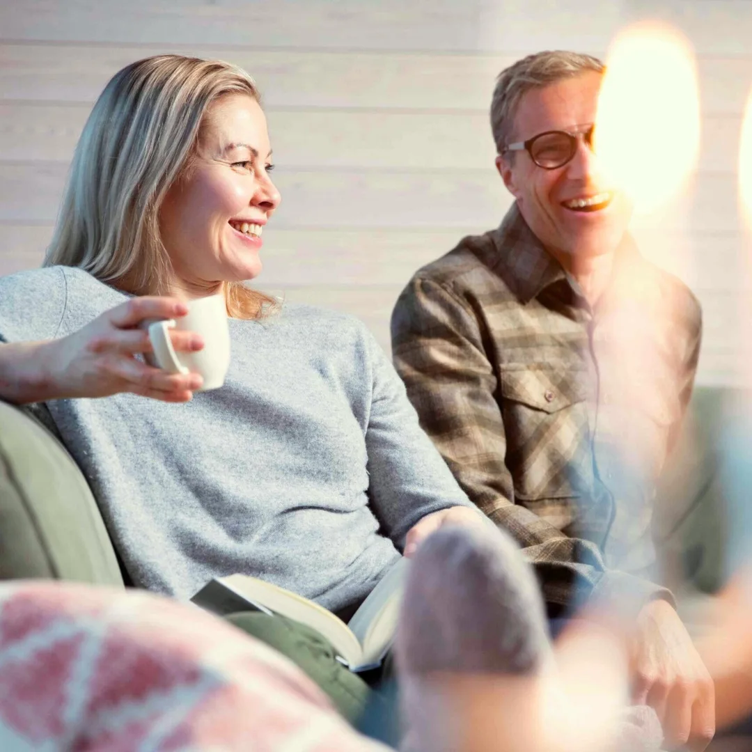 Nainen ja mies nauravat sohvalla kotoisassa tunnelmassa Pohjois-Karjalassa, naisella kahvikuppi kädessä – vastuullista matkailua kotimaan matkailua.