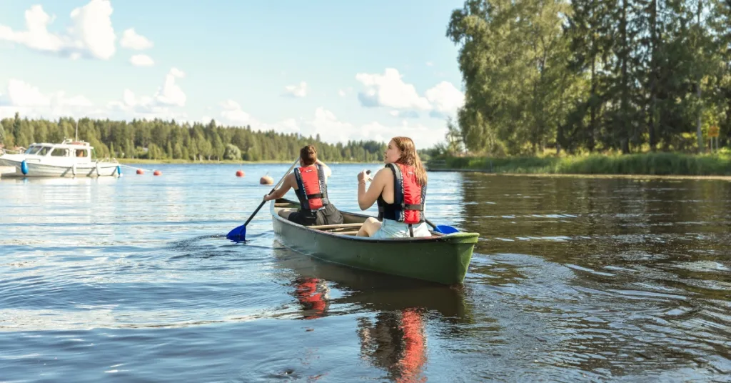 Kaksi tyttöä meloo vihreässä intiaanikanootissa Pohjois-Karjalassa – kauempana satama ja lähellä metsäinen ranta.