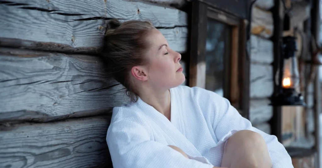 Nainen nojaa hirsiseinään silmät kiinni saunan jälkeen Pohjois-Karjalassa – rentoutumista vuokramökeissä ja leirintäalueilla.