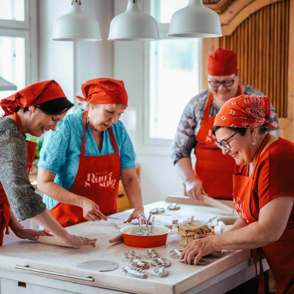 Joukko naisia punaisissa esiliinoissa leipoo karjalanpiirakoita iloisissa tunnelmissa Pohjois-Karjalassa – koe karjalanpiirakka-pajat Ohjelmapalvelut-sivulta.