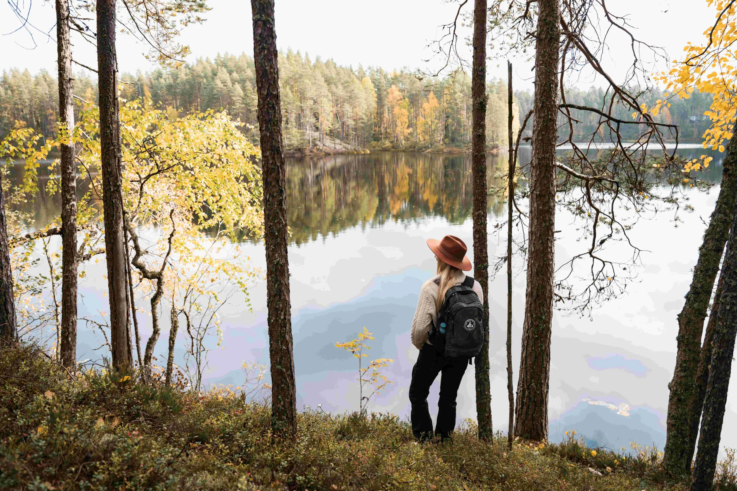Nuori nainen lierihattu päässään katsoo syksyiselle järvelle takaapäin Pohjois-Karjalassa – rauhallisia patikointielämyksiä luonnossa.